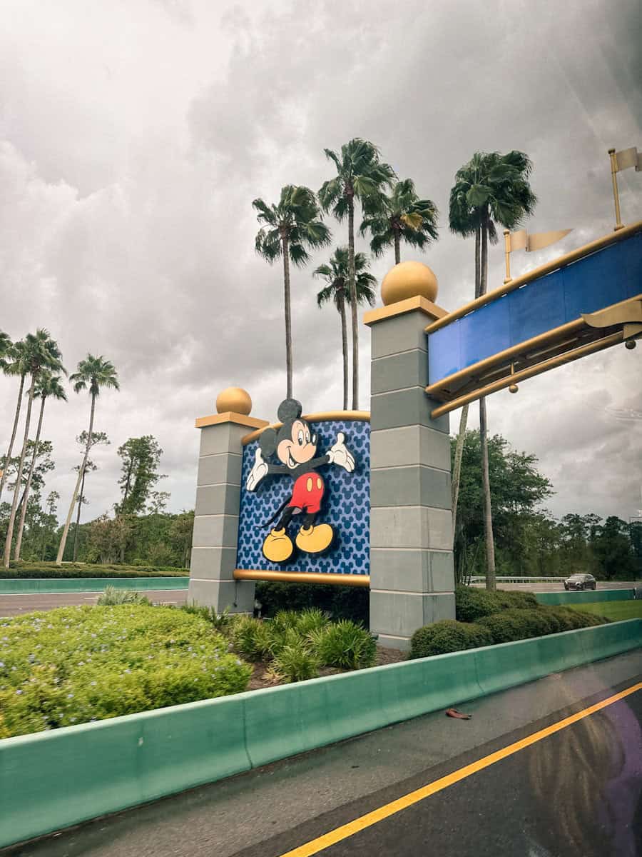 Driving on the bus to Disney World in the rain, looking out the window to the welcome sign with Mickey Mouse and dark clouds in the background.